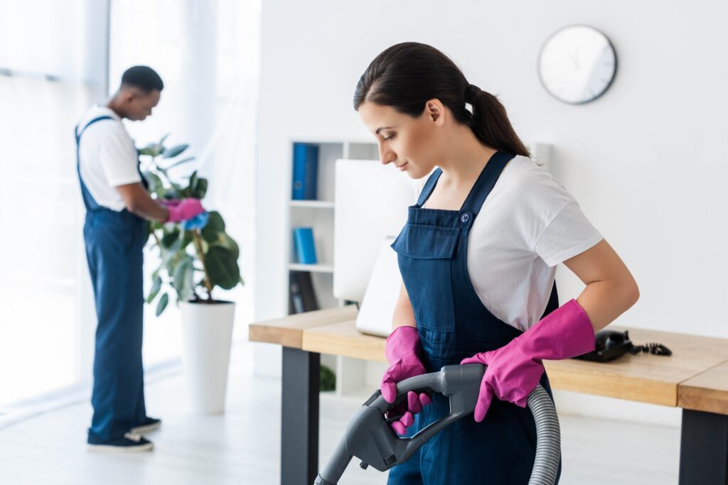 Selective focus of attractive worker of cleaning service using vacuum cleaner near african american
