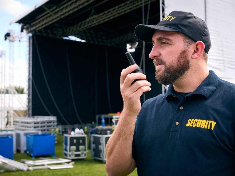 Security Team At Outdoor Stage For Music Festival Or Concert Talking Into Radio