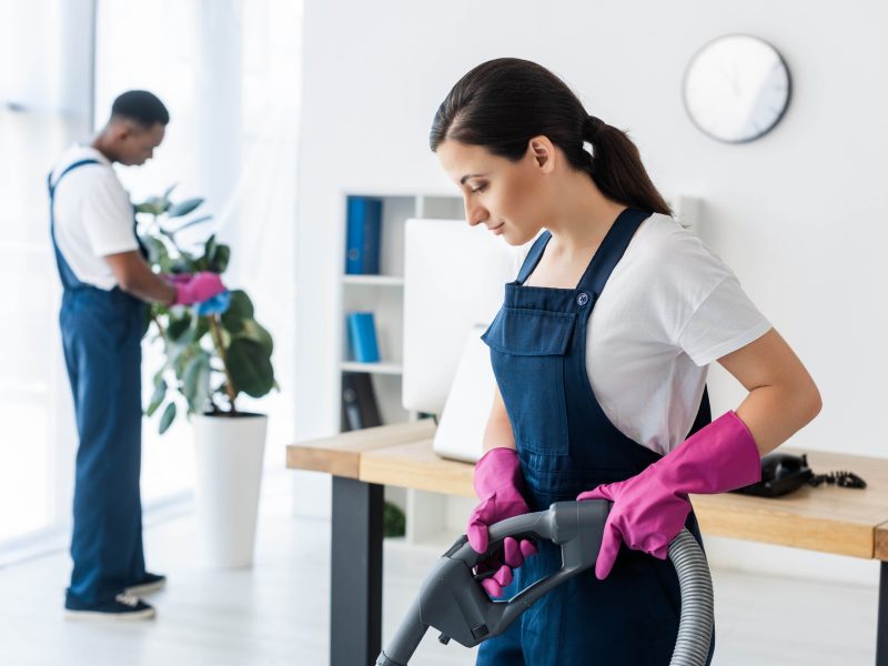 Selective focus of attractive worker of cleaning service using vacuum cleaner near african american