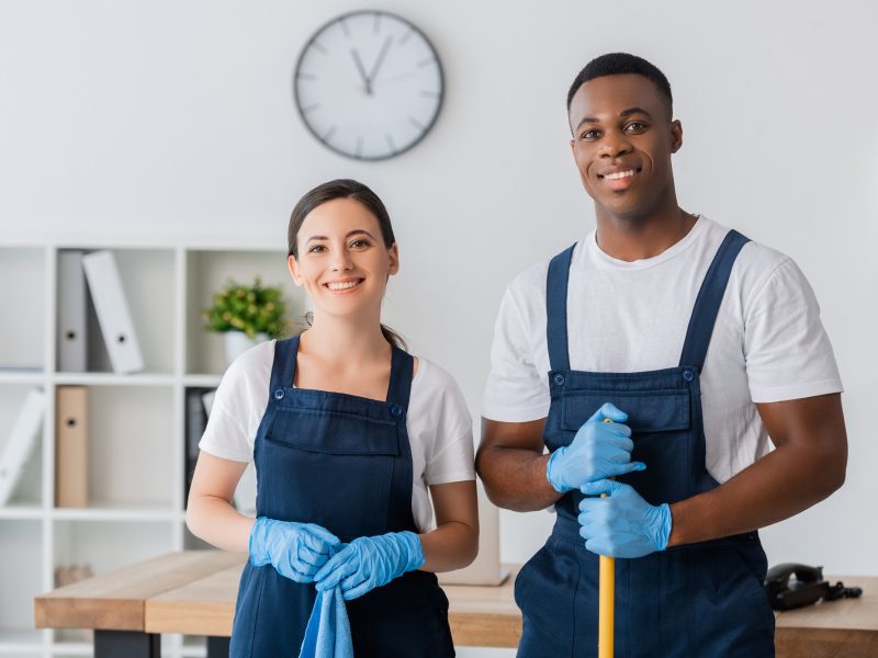 Smiling multiethnic workers of cleaning service holding rag and mop while working in office